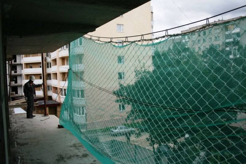 A building under construction, its balcony with green fabric sheet and a chain link fence reinforcement.