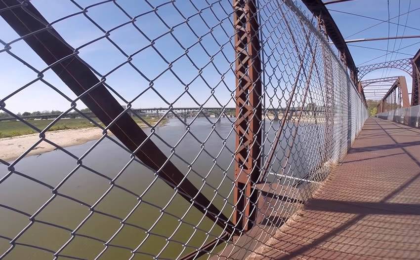 A metal bridge with chain link railing across a deep and wide river.