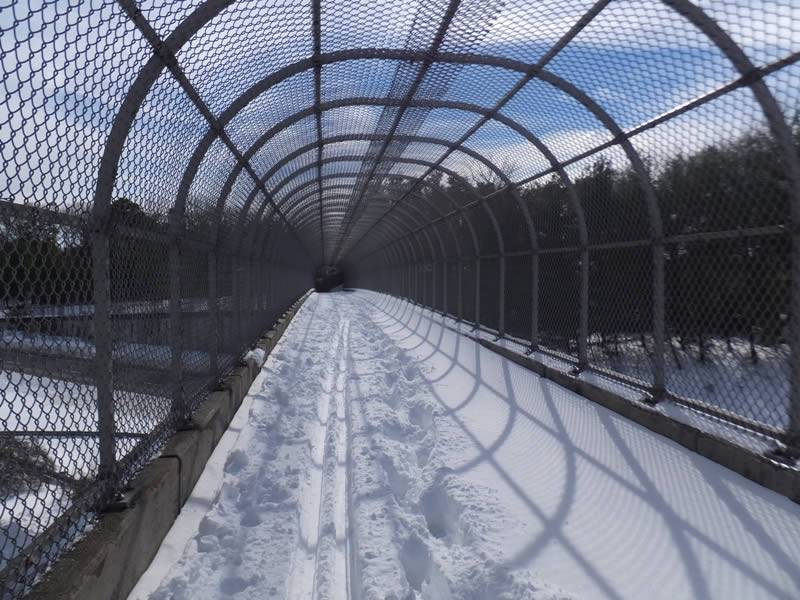 Walkway in ski resort with semicircle chain link roof, and the walkway covered by snow.